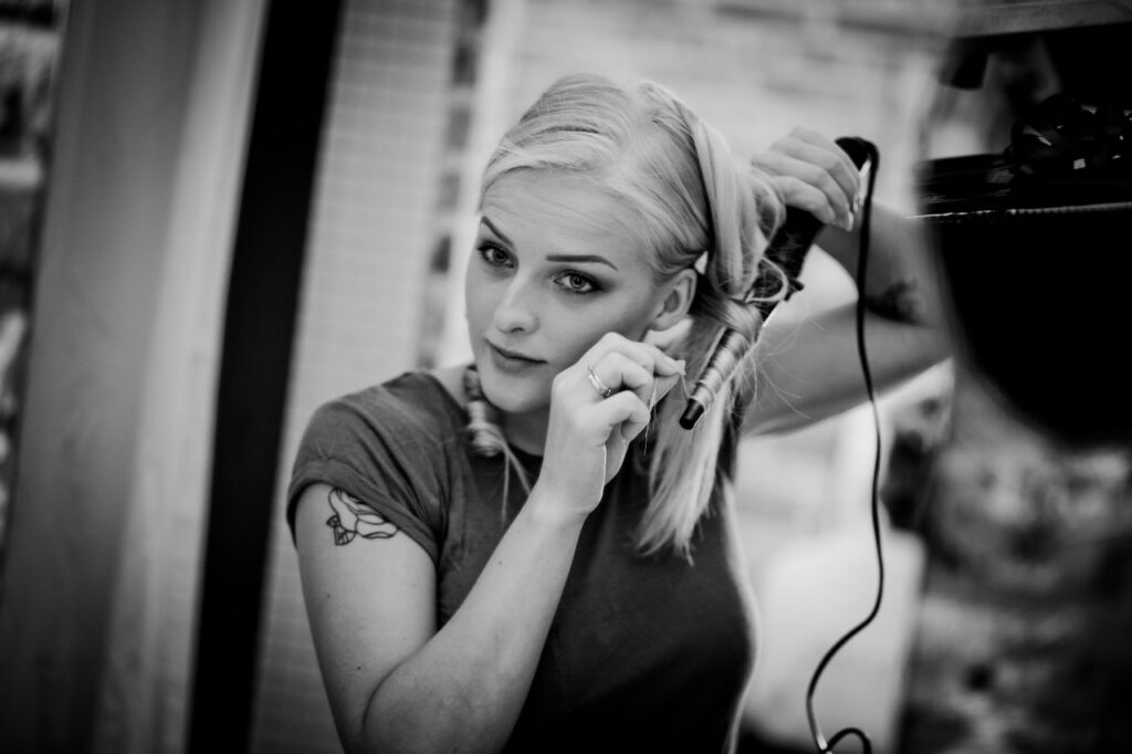 A young woman with tattoos using a curling iron to style her hair in a modern indoor setting.