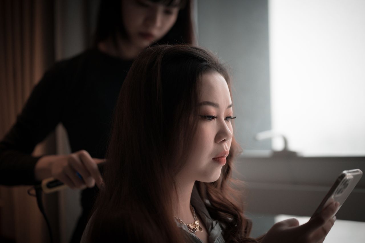 An Asian woman using her phone while a hairstylist styles her hair indoors.